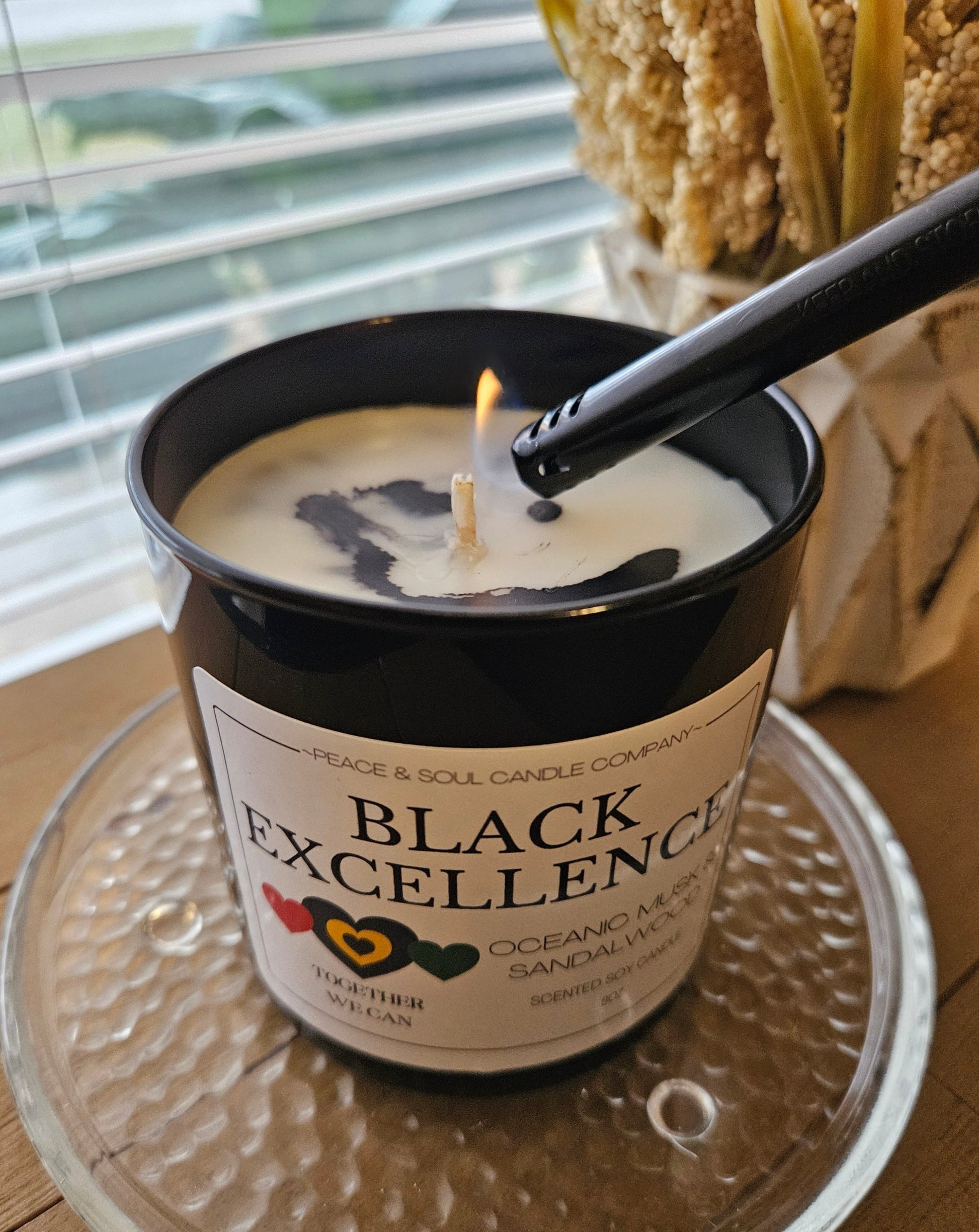 black candle for black history month in black glass jar with white label decorated with red, green and yellow hearts. Lighter lighting the candle. candle is sitting on a desk with a view of the outside.